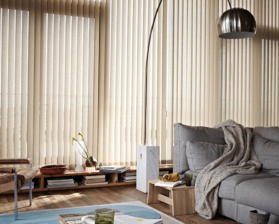 Beige verticale lamellen groot raam Luxaflex verticale jaloezieën grijze bank houten vloer